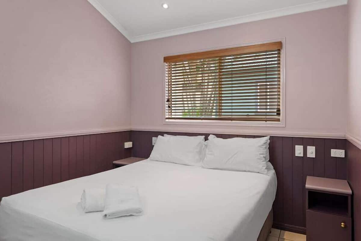 A neatly made bed with white linens and a folded towel, a window with horizontal blinds, and a small bedside table with a lamp.
