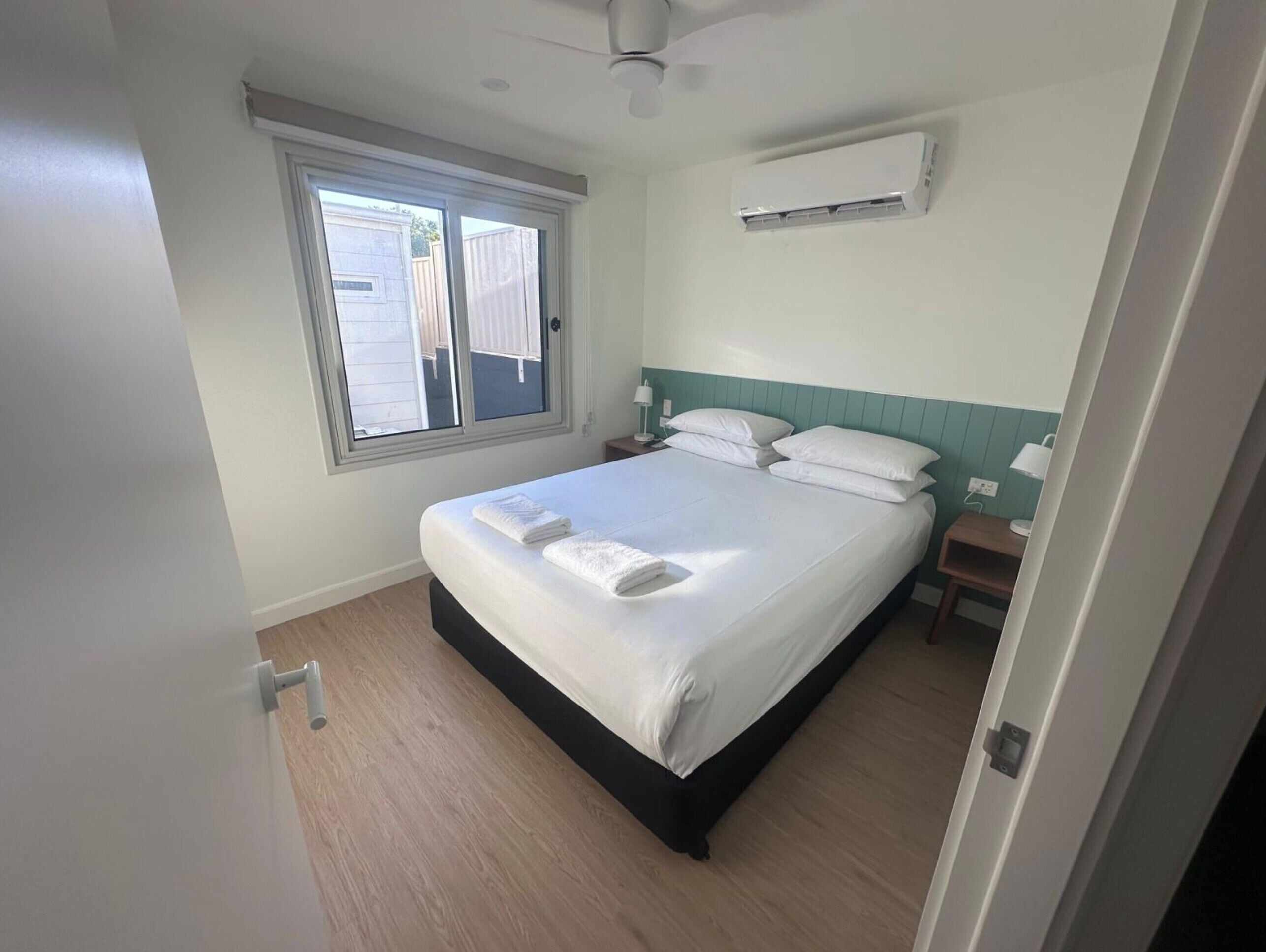A bedroom with a bed, a window, an air conditioning unit, a nightstand, and a bedside table.