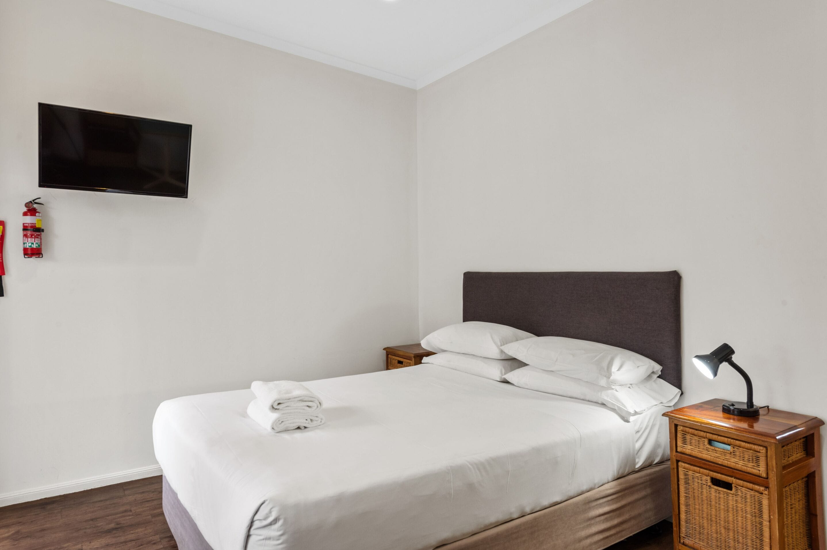 Una cama bien hecha con ropa blanca, un televisor montado en la pared, una mesita de noche con una lámpara y un extintor en la pared.