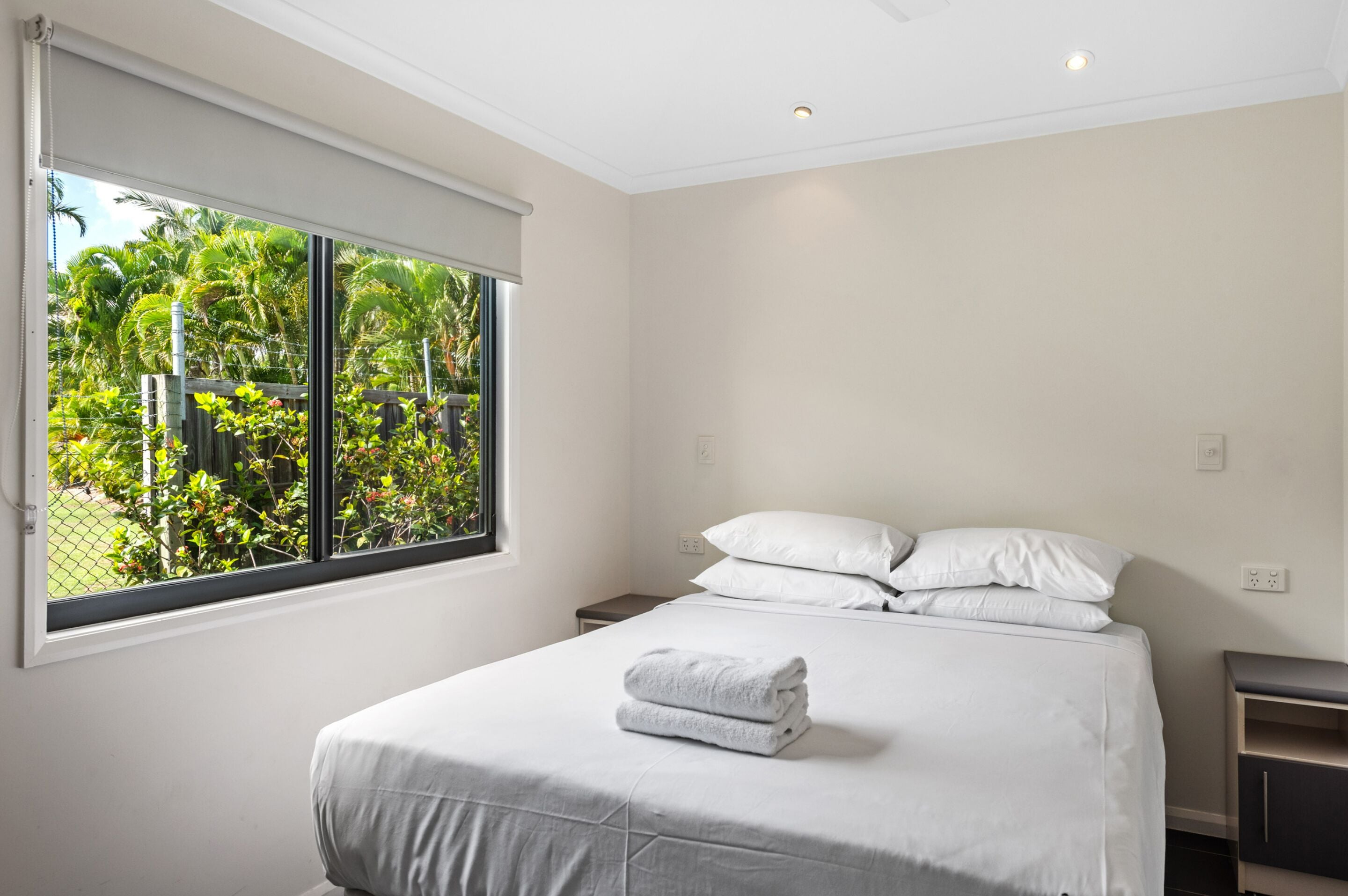 A bedroom with a bed, pillows, a nightstand, a window with a view of greenery, and a ceiling light.