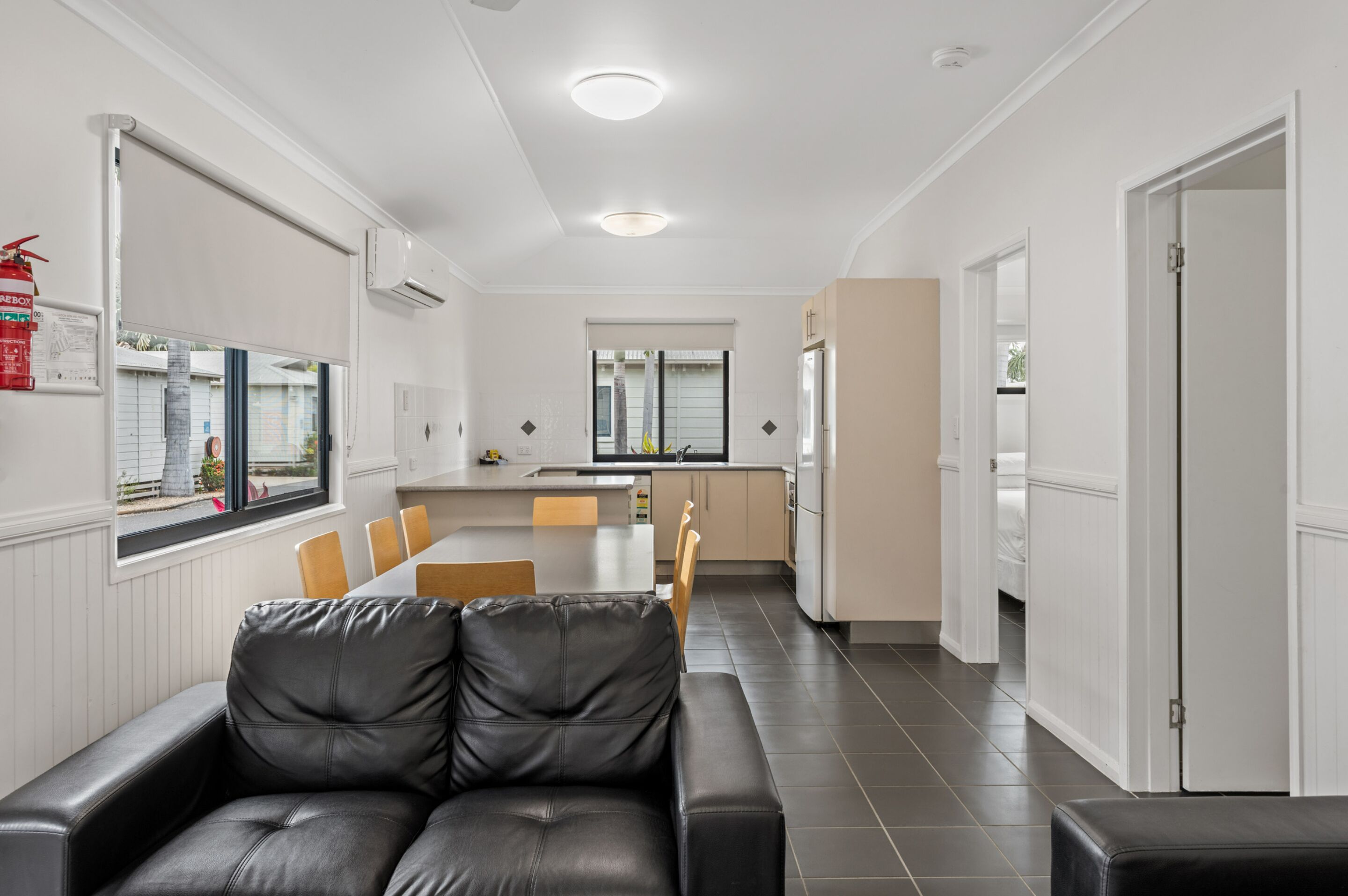 A modern kitchen with white cabinets, a central island, and a dining area with a black leather sofa and wooden chairs.