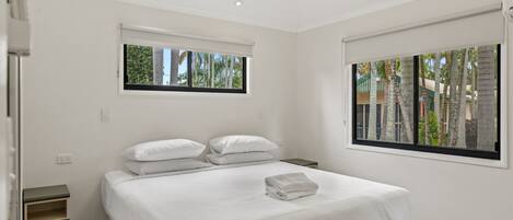 Une chambre d’hôtel moderne avec un grand lit, deux fenêtres, un ventilateur de plafond et une petite table de chevet.