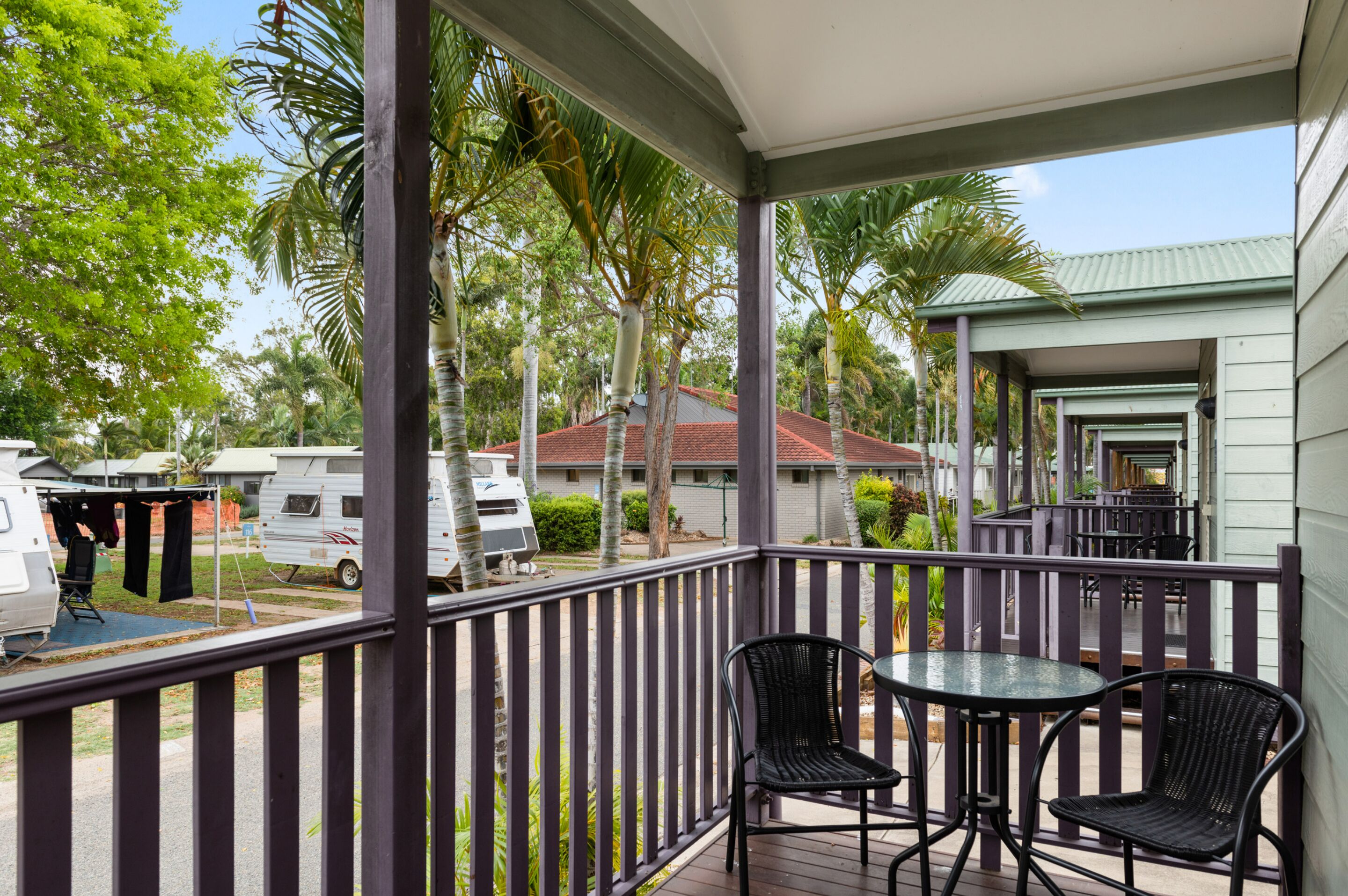 A covered outdoor area with a table and chairs, surrounded by a railing, overlooking a residential area with houses and a parked RV.