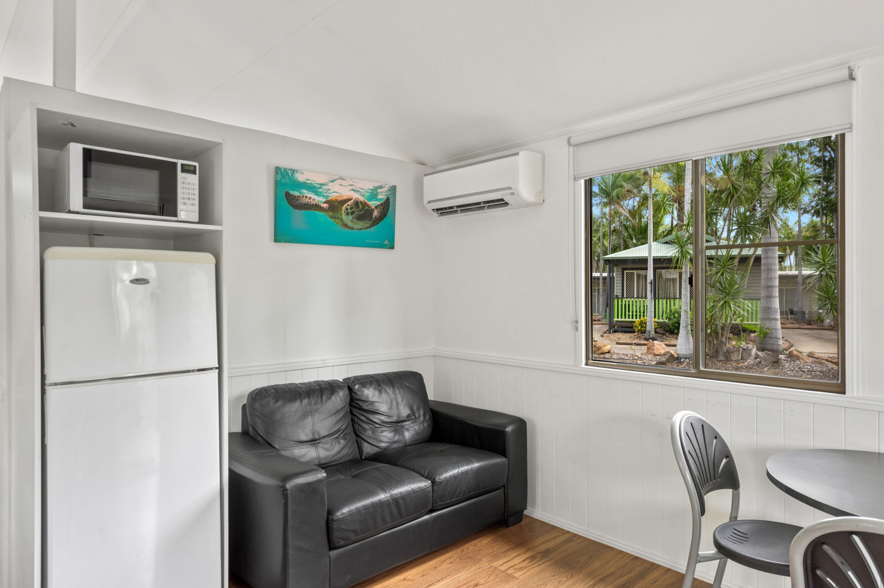 A compact room with a white refrigerator, a black leather sofa, a dining table with two chairs, a microwave, and a painting of a sea turtle.