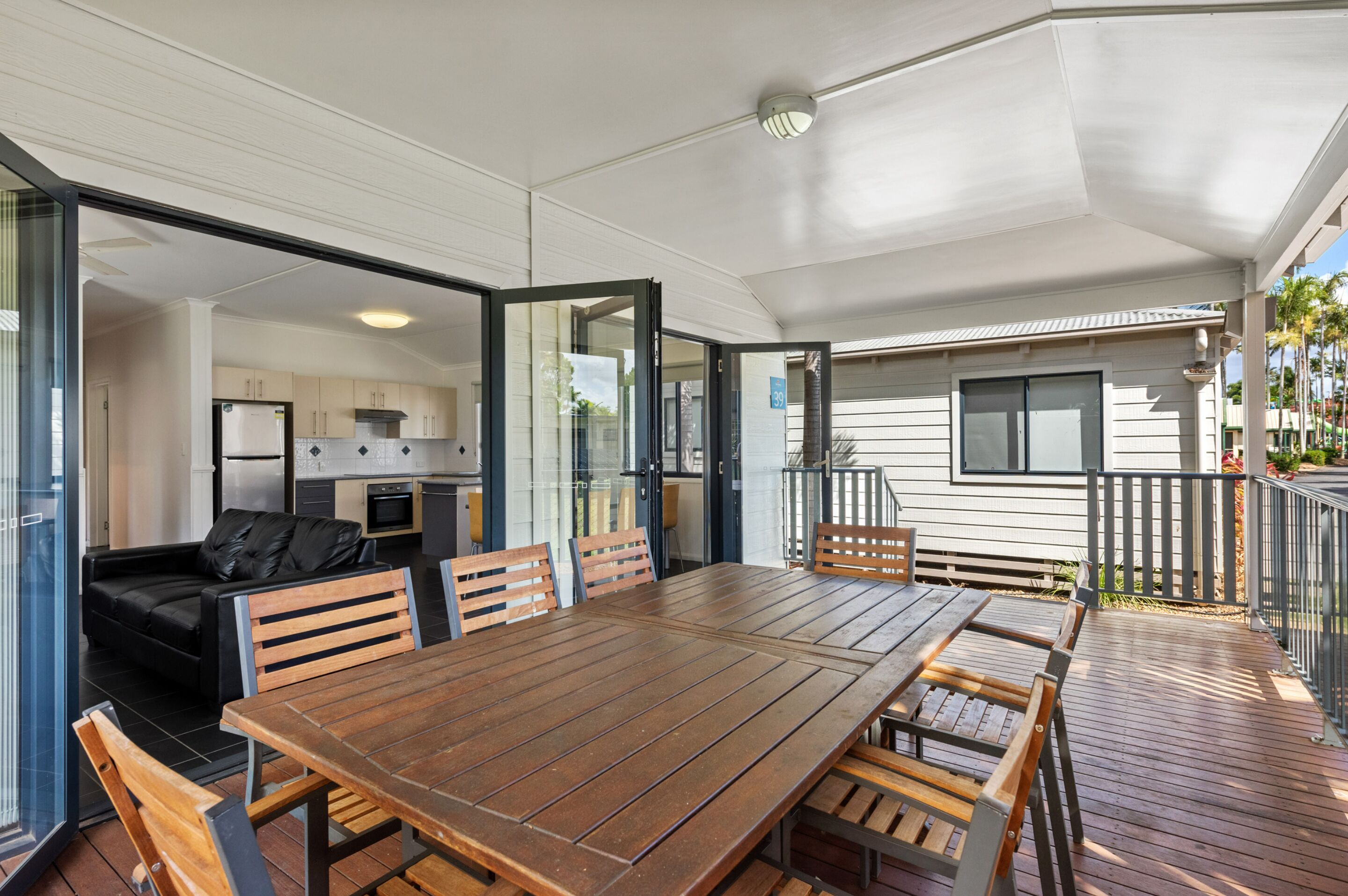 A spacious outdoor dining area with a wooden table and chairs, a black leather sofa, and a glass door leading to a kitchen.