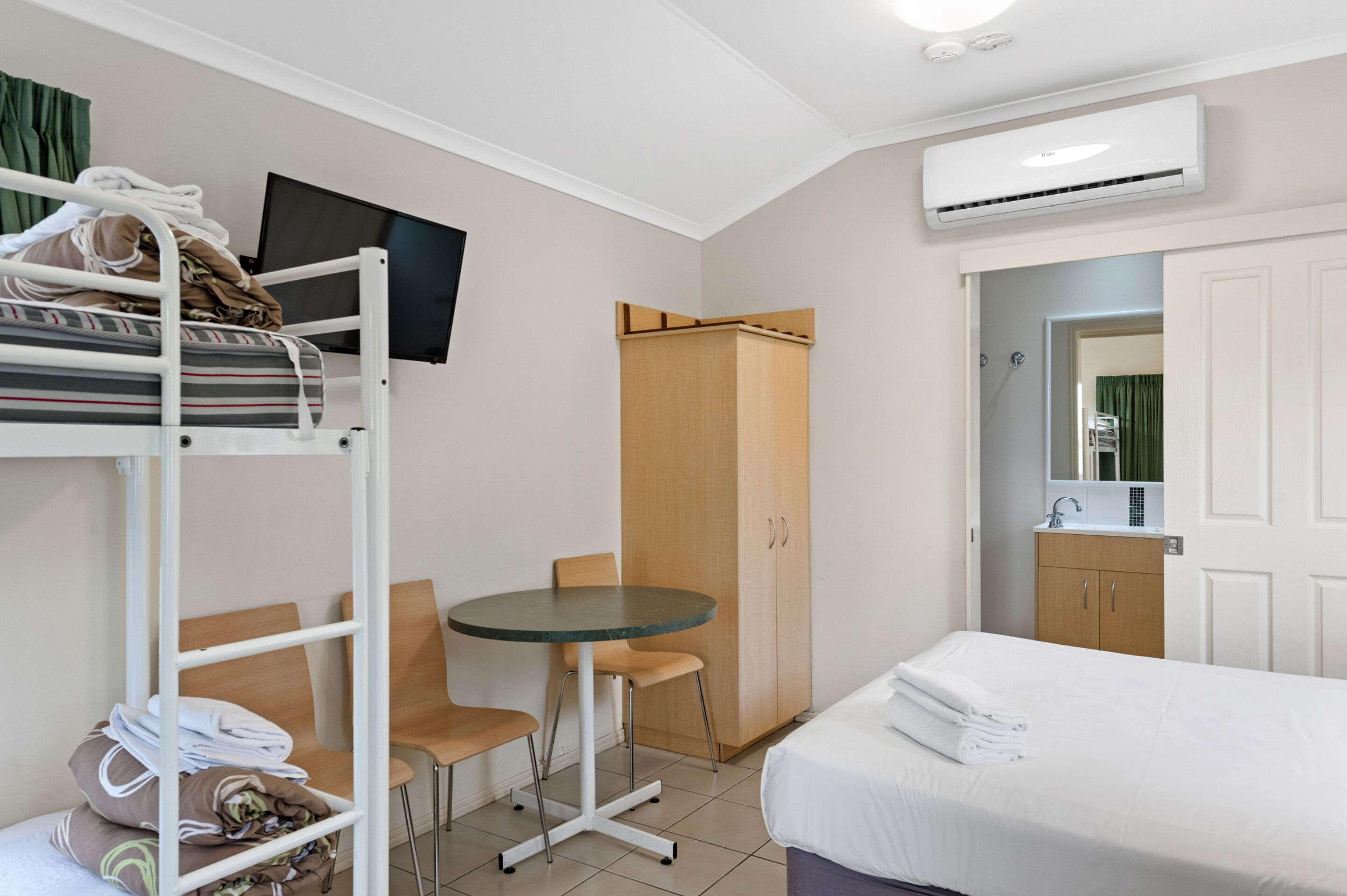 Ein Hotelzimmer mit einem Etagenbett, einem kleinen Tisch mit Stühlen, einem Kleiderschrank, einem Fernseher und einem durchgegangenen Tür sichtbaren Badezimmer.