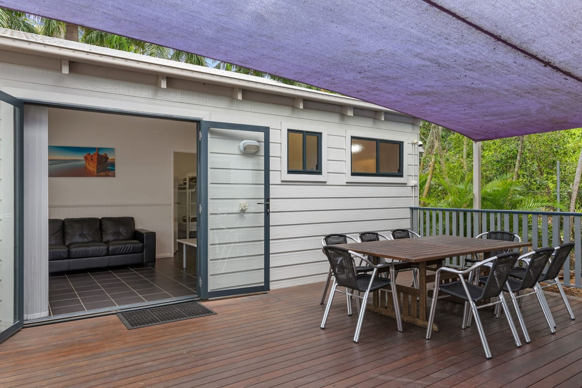 A covered outdoor area with a wooden deck, a dining table with chairs, a sofa, and a small building with windows and a door.