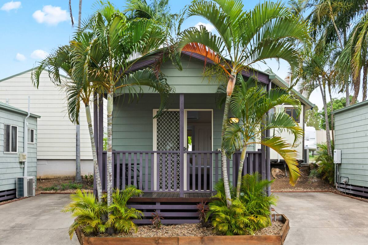 A small, single-story house with a porch, surrounded by palm trees and a concrete driveway.
