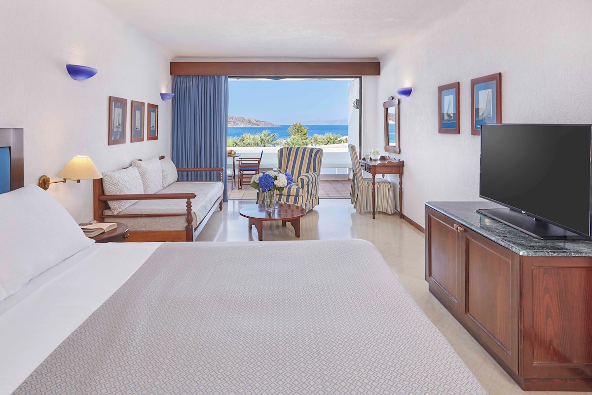 Habitación de hotel con una cama grande, televisión, escritorio y balcón con vista al mar.