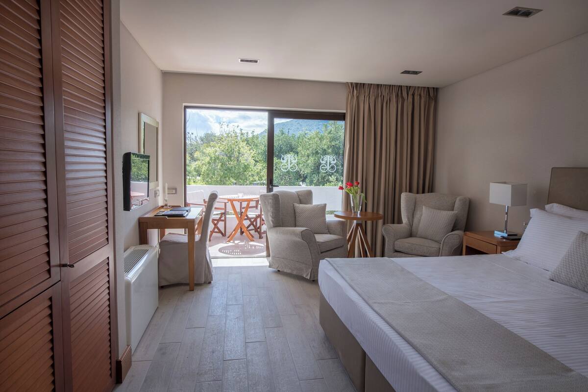 Habitación de hotel con cama, dos sillones, un escritorio y una mesa. Se ve una ventana con vistas a árboles y montañas.