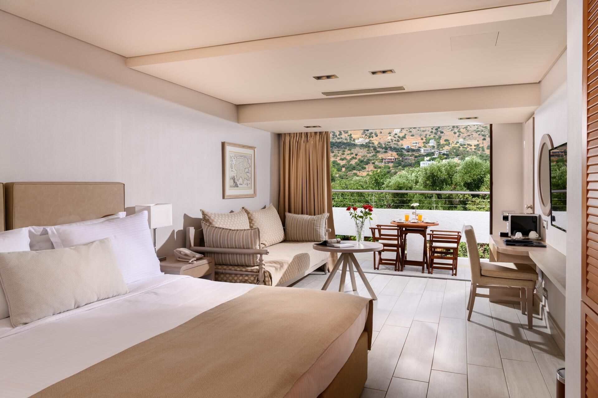 Ein Hotelzimmer mit Bett, einer Sitzecke, einem Esstisch mit Stühlen, einem Fernseher und einem Balkon mit Blick auf die Landschaft.