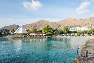 Una piscina azul cristalina con un velero, sombrillas de playa y un hotel con las montañas al fondo.