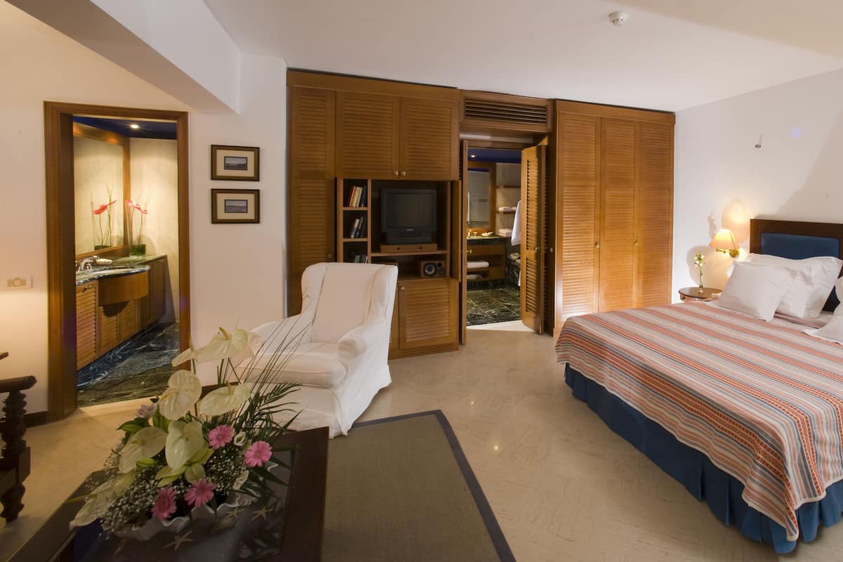 Habitación de hotel con cama, una silla, un televisor y un baño visible a través de una puerta abierta.