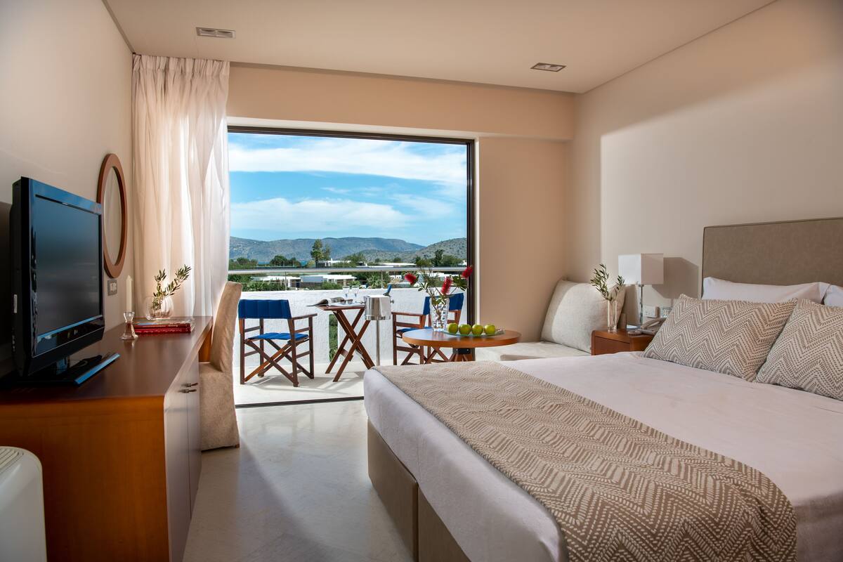 Habitación de hotel con una cama grande, televisión, escritorio con computadora, balcón con mesa y sillas, y vistas a las montañas.