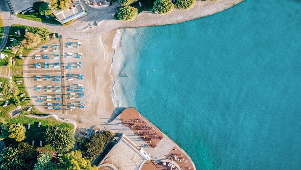 An aerial view of a coastal area with a beach, a parking lot, and a building with a red roof. - Elounda Bay Palace, a Member of the Leading Hotels of the World (Agios Nikolaos)
