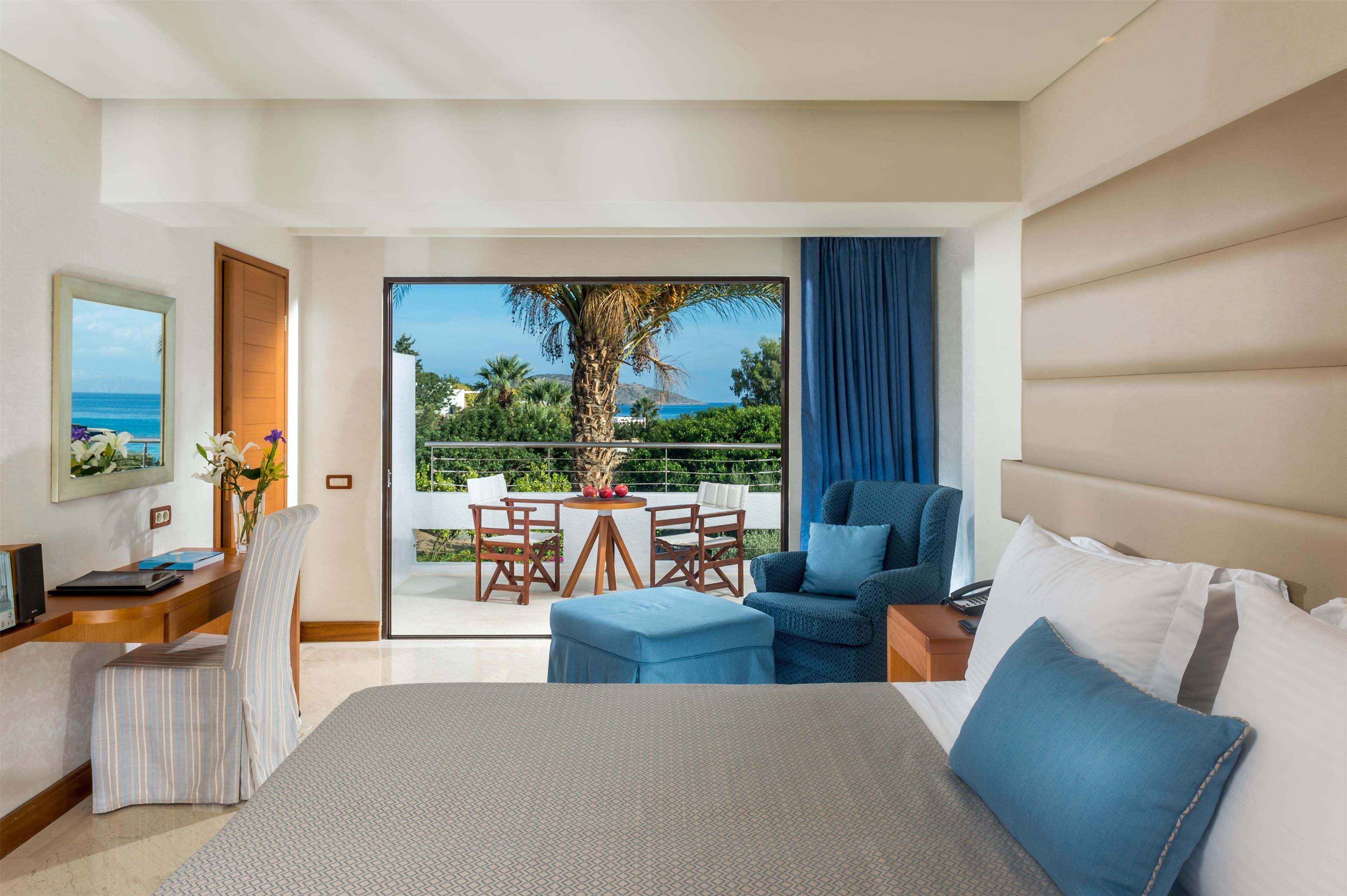 Una habitación de hotel con una cama grande, un escritorio con silla, una mesita con sillas y vistas al océano a través de amplias ventanas.