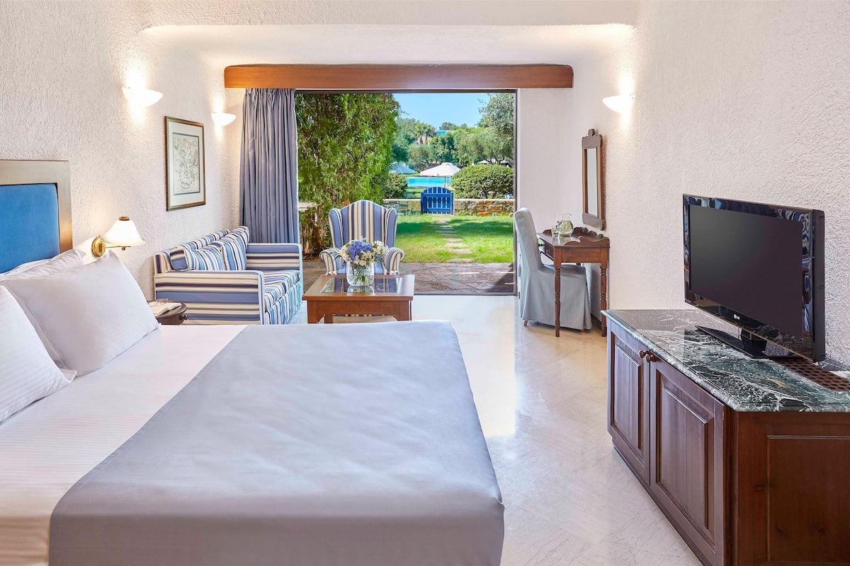 Habitación de hotel con una cama grande, un sofá, un televisor y un escritorio. Se ve un jardín desde la puerta.