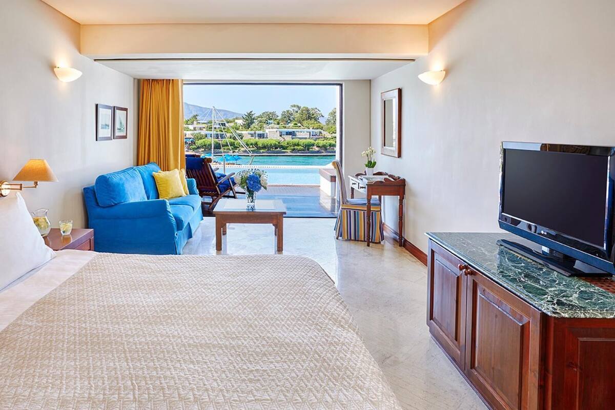 Habitación de hotel con cama, sofá azul, televisor, escritorio y vistas a una piscina y montañas.
