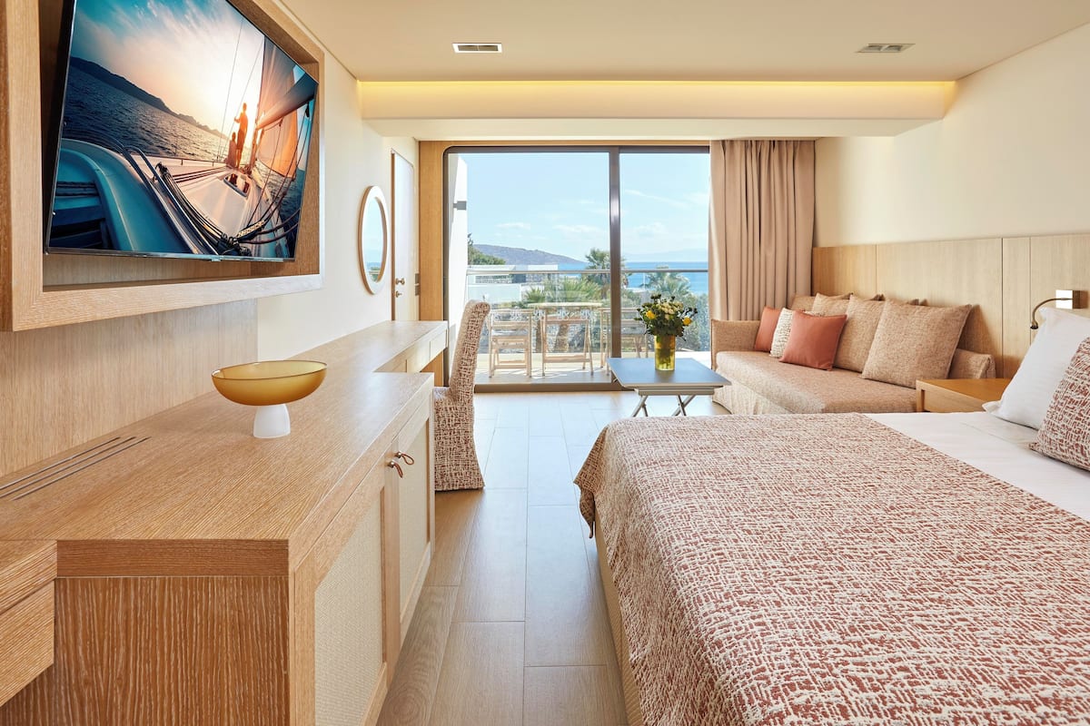 Habitación de hotel con televisor grande, cama, sofá, mesa de comedor y vista al mar.