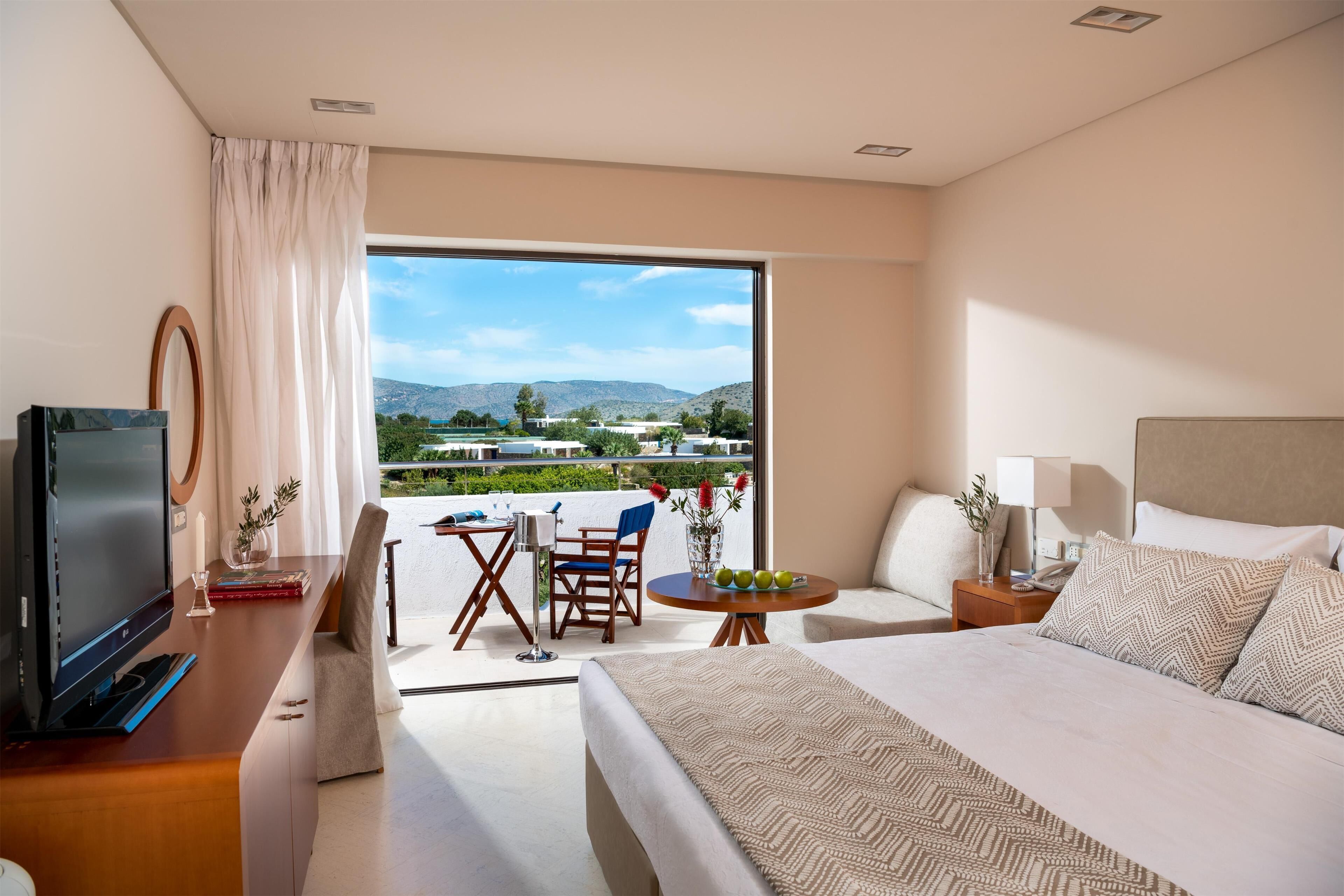 A modern hotel room with a large bed, a desk with a computer, a chair, a small table with fruit, and a view of the outdoors.