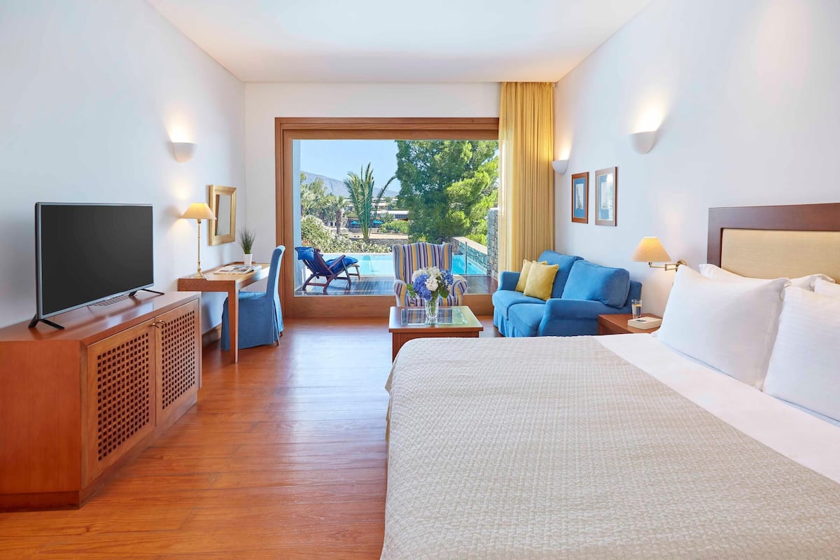 Habitación de hotel con una cama grande, televisión, sofá, sillón, una mesita y vistas a una piscina y palmeras.