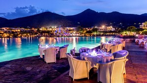 A waterfront restaurant with tables and chairs set up for dining, overlooking a pool and mountains in the background. - Elounda Bay Palace, a Member of the Leading Hotels of the World (Agios Nikolaos)