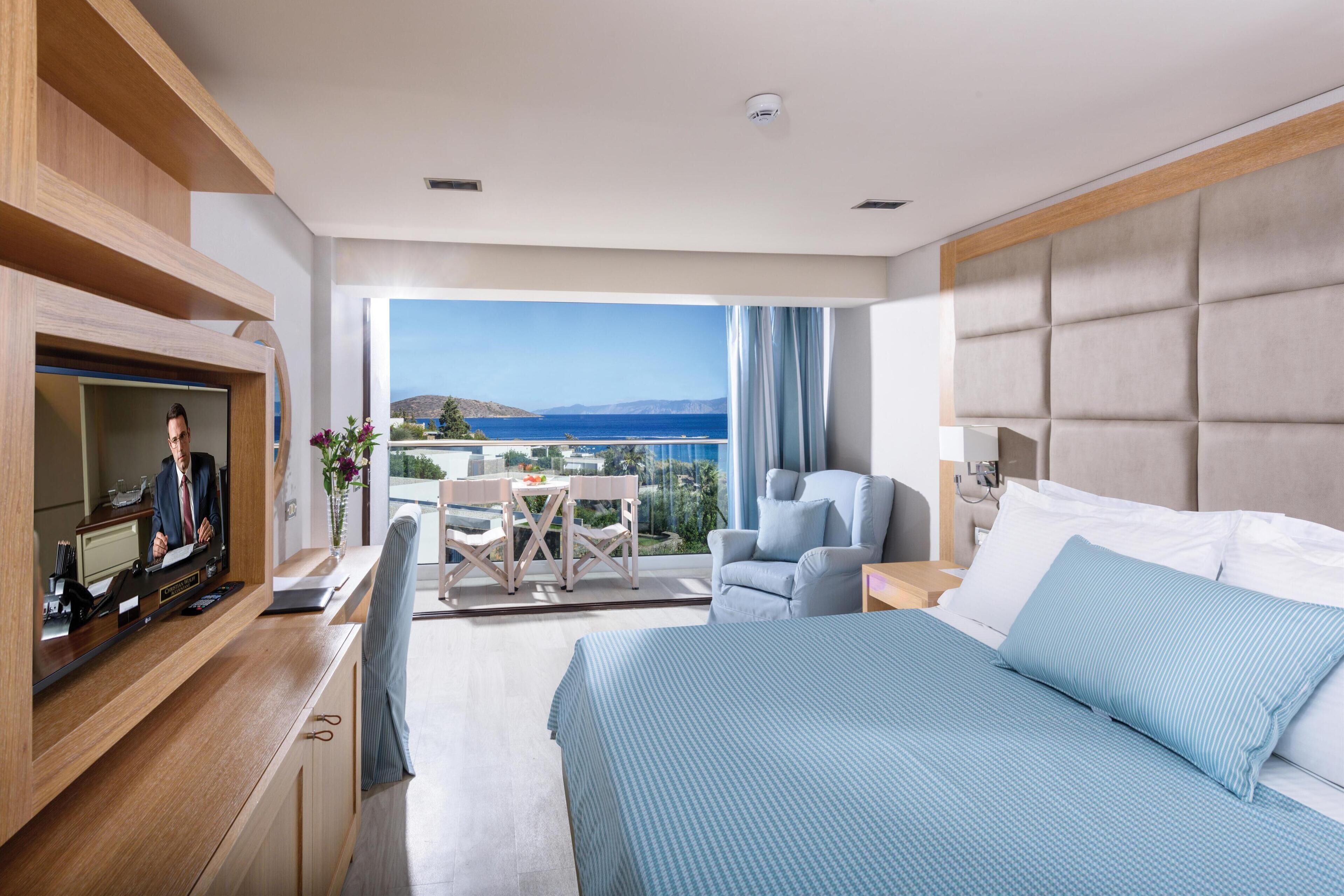 A modern hotel room with a large bed, a TV, a desk, and a balcony with a view of the sea.