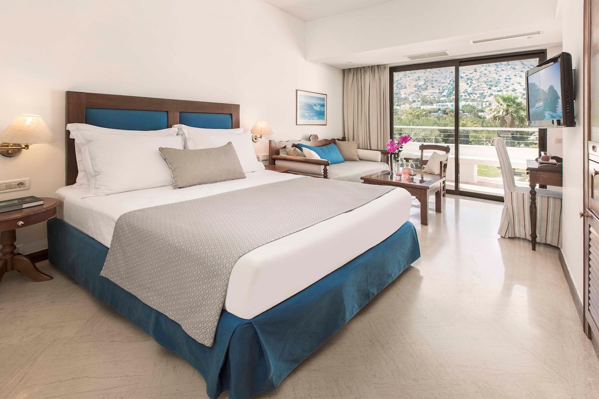 Habitación de hotel con una cama grande, un sofá, un televisor y un balcón con vistas al paisaje.