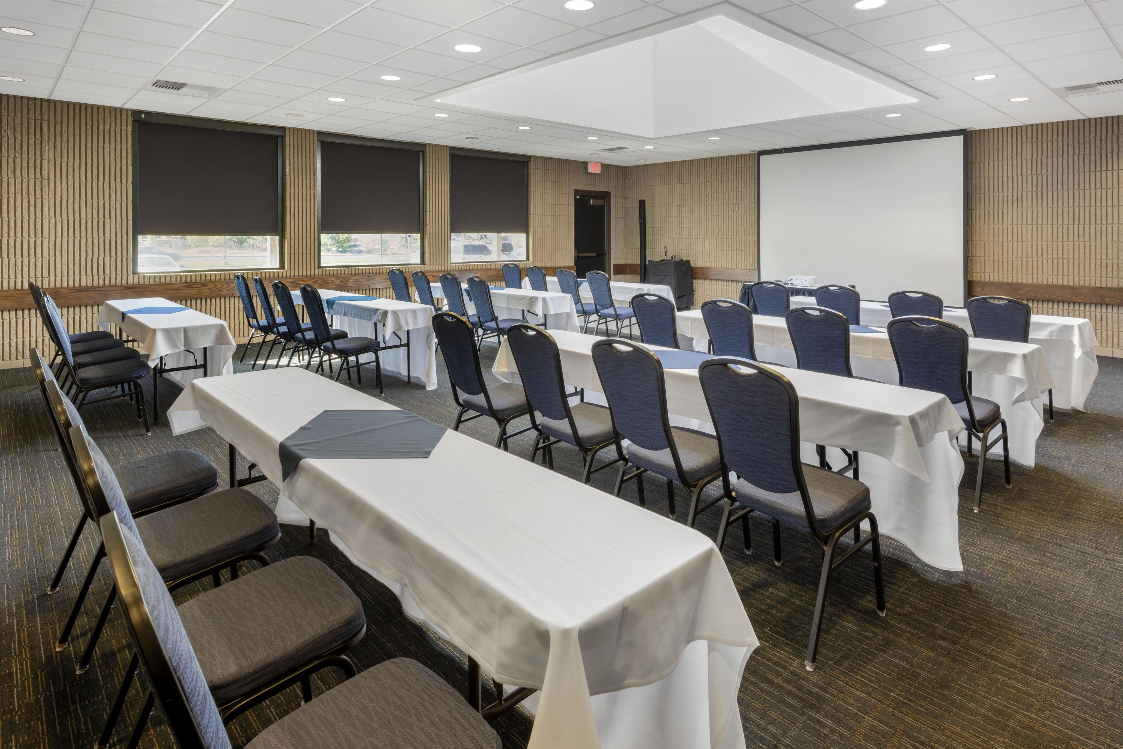 Sala de conferências com várias mesas e cadeiras, uma tela de projetor e uma janela grande.