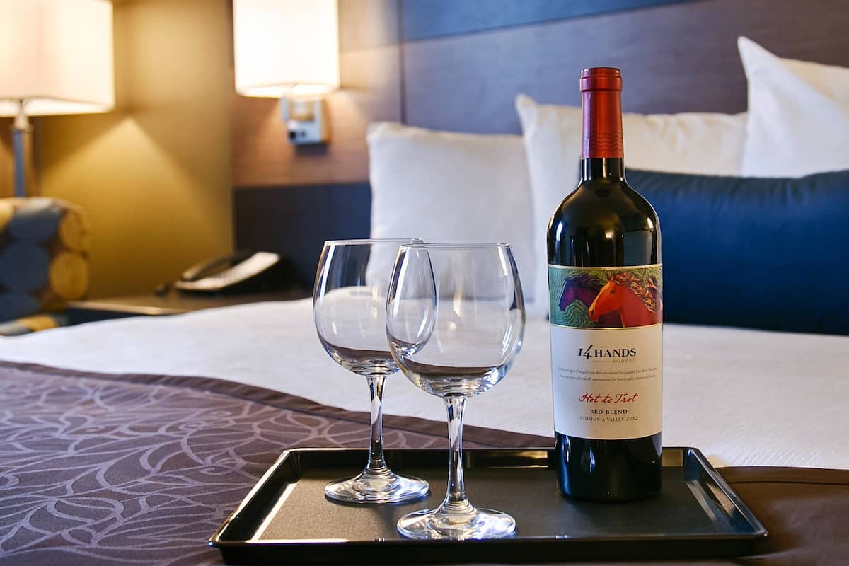 A hotel room with a bed, two bedside tables, a lamp, a phone, and a tray with a wine bottle and two glasses.