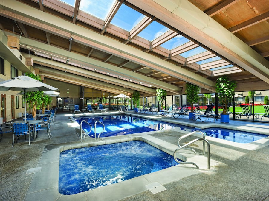 An indoor swimming pool with multiple lanes, lounge chairs, and tables under a large, open roof structure.