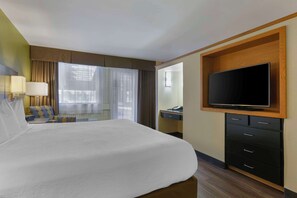 A hotel room with a large bed, a television mounted on a wooden panel, a desk with a chair, and a window with curtains.