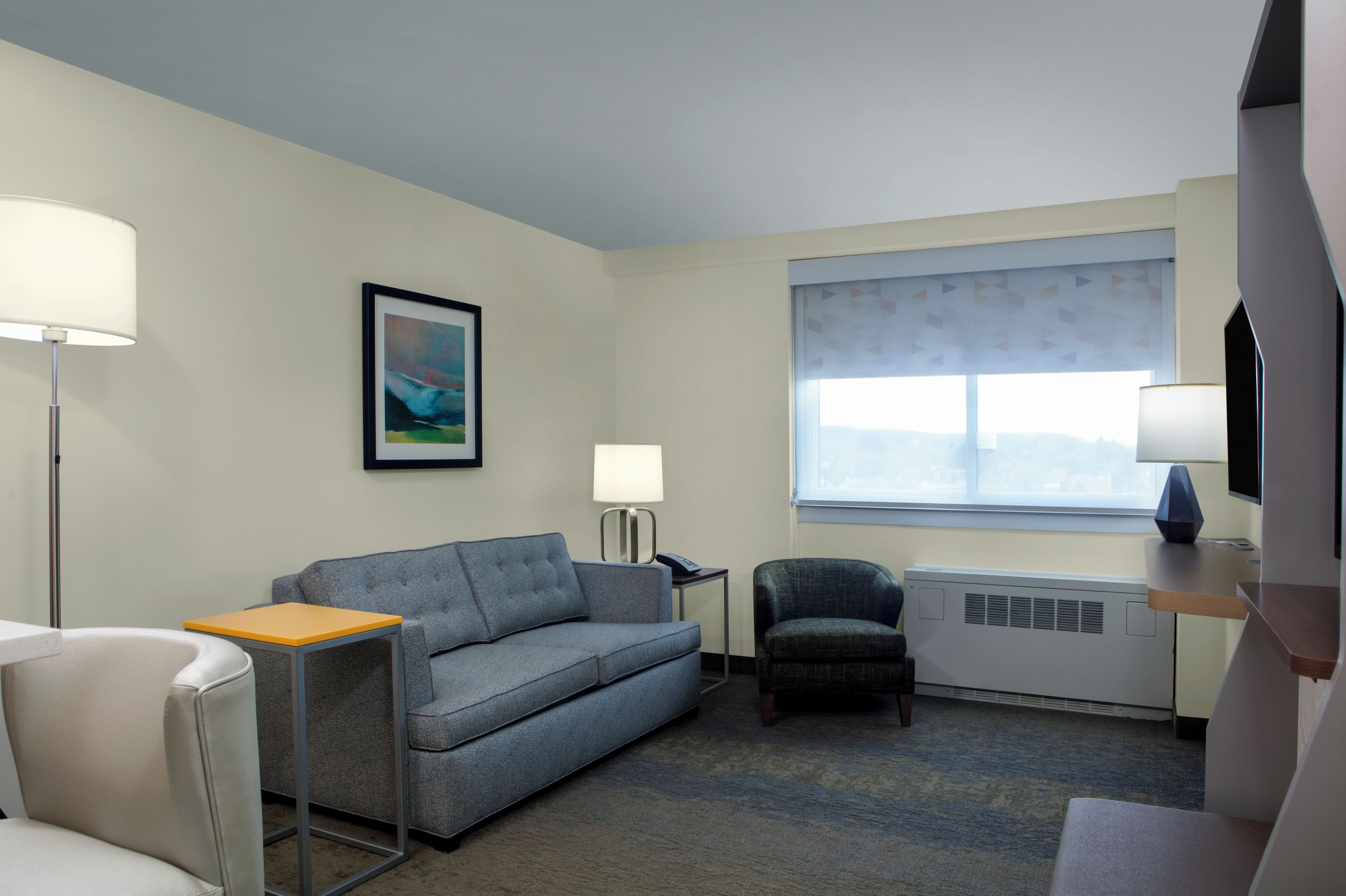 A hotel room with a grey sofa, a small table, a chair, a standing lamp, a framed picture on the wall, a window with a view, and a TV mounted on the wall.