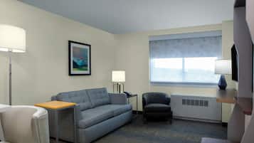 A hotel room with a grey sofa, a small table, a chair, a standing lamp, a framed picture on the wall, a window with a view, and a TV mounted on the wall.