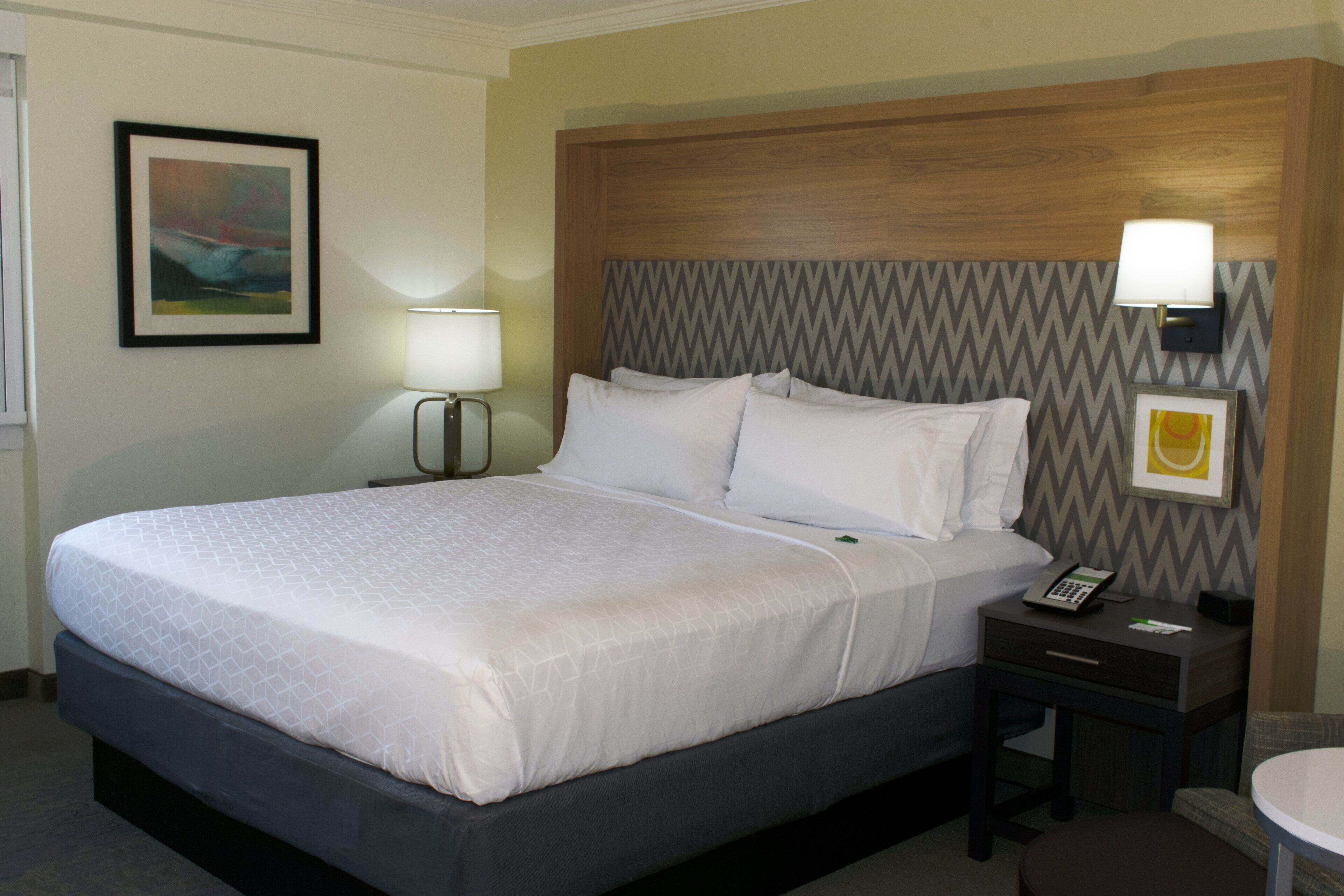 A neatly made bed with a white comforter and pillows, a bedside table with a lamp, and a framed picture on the wall.