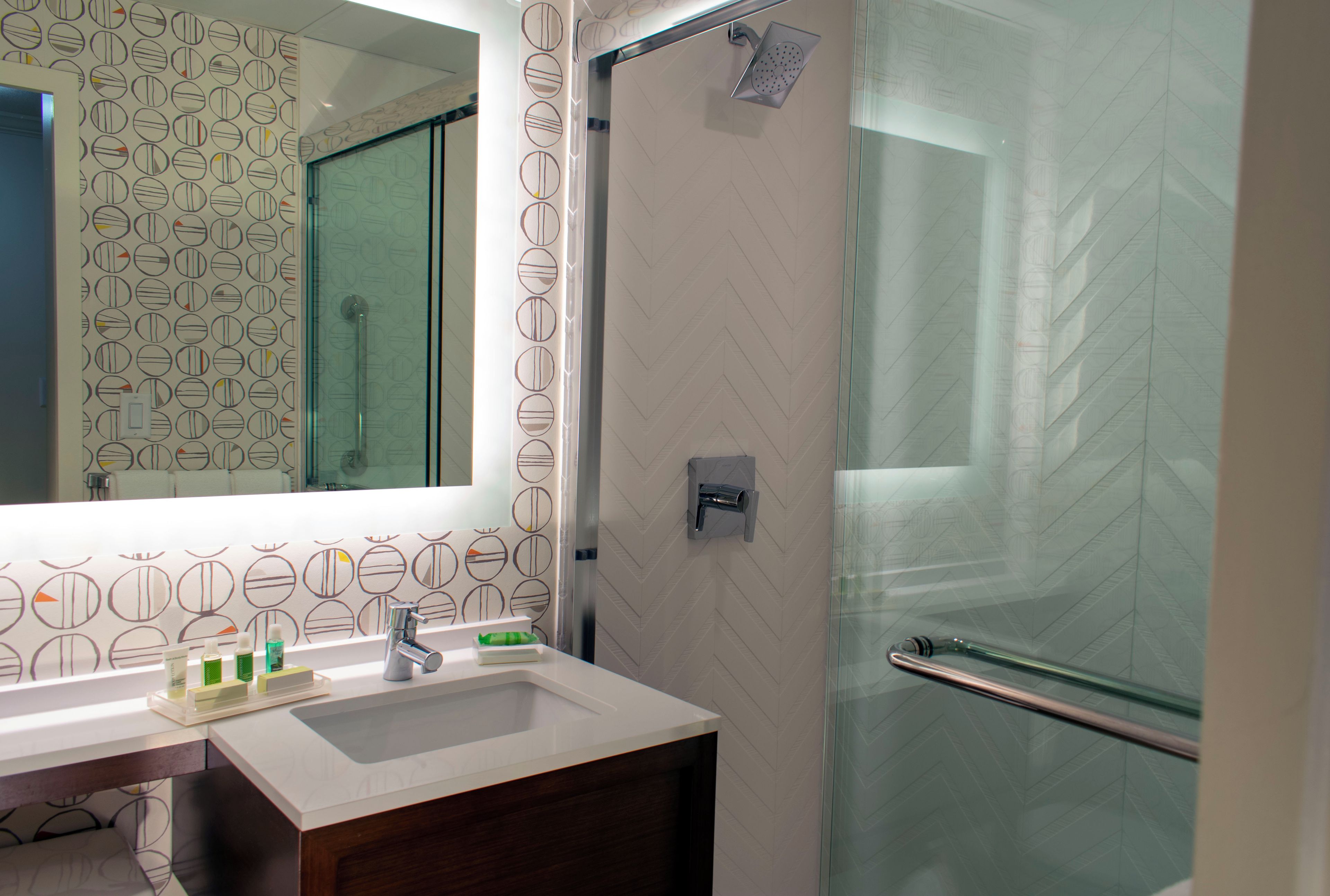 A modern bathroom with a glass shower enclosure, a white sink, and a mirror with a circular pattern wallpaper.