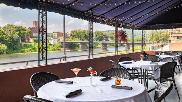 Un espace extérieur aménagé pour les repas, avec des tables et des chaises, offrant une vue sur une rivière et des bâtiments.