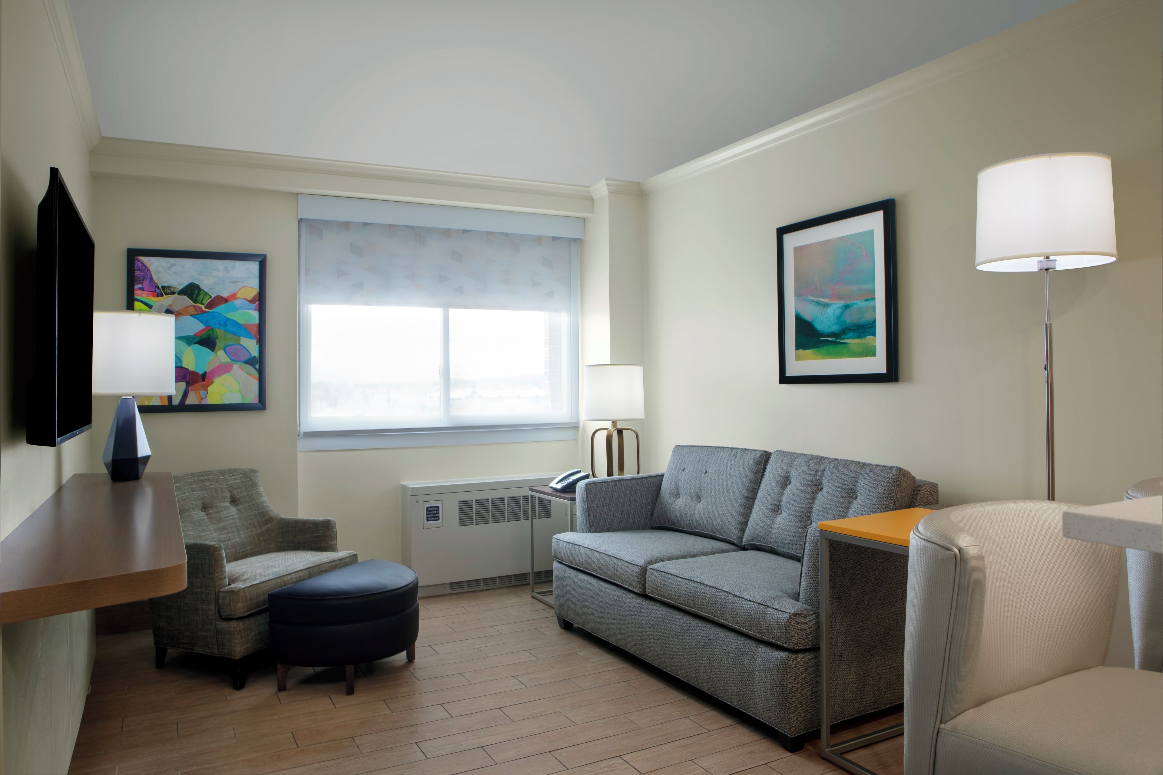 A modern hotel room with a grey sofa, a small round table, a flat-screen TV, a window with a view, and a painting on the wall.
