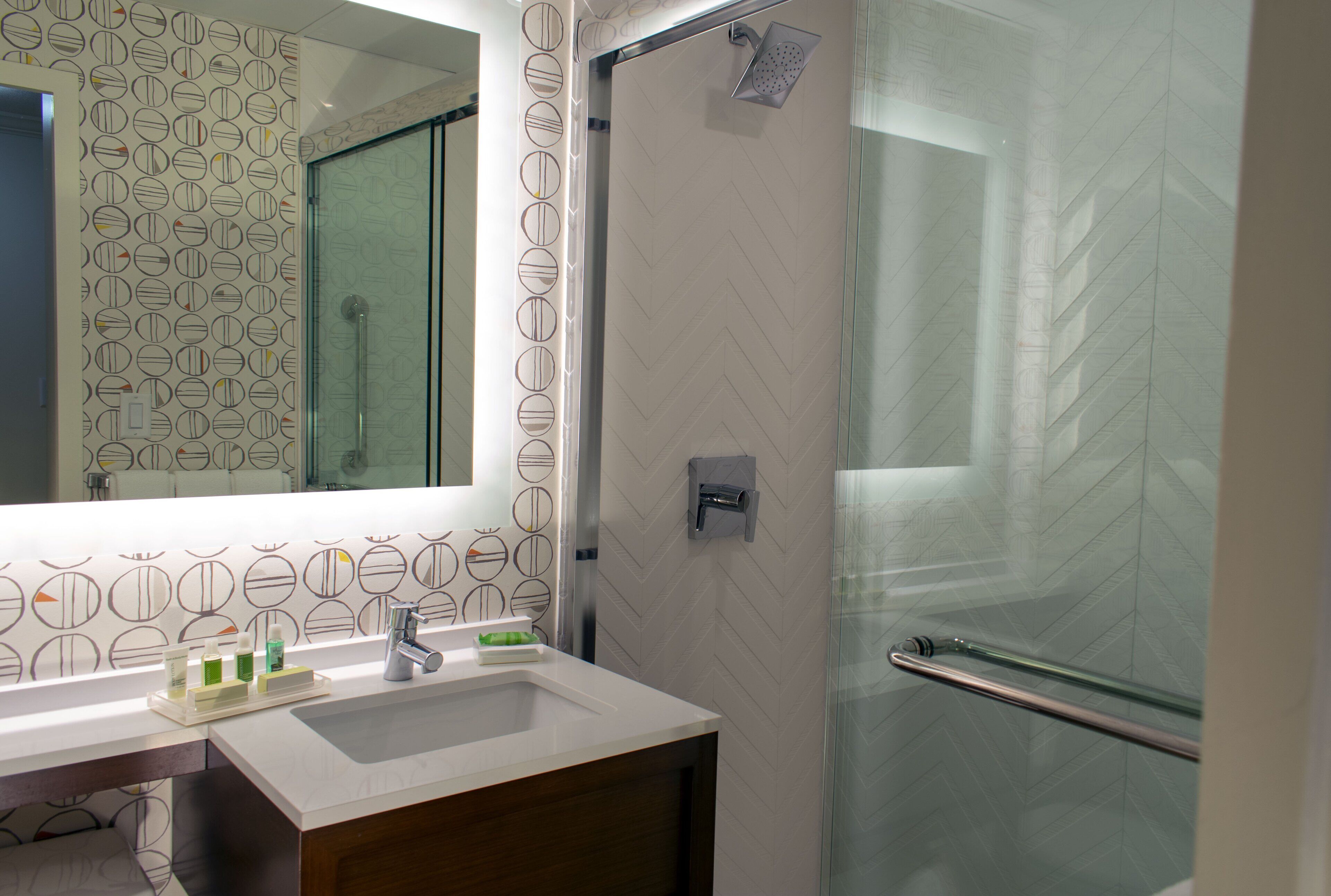 A modern bathroom with a glass shower enclosure, a white sink with a mirror above it, and patterned wall tiles.