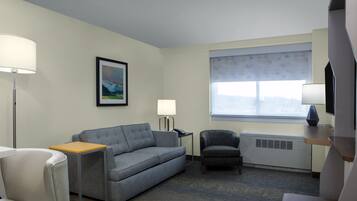 Habitación de hotel con un sofá gris, una mesita, una silla, una lámpara de pie, un cuadro en la pared y un gran ventanal con vistas.