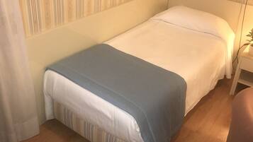 A single bed with a white and blue bedspread, a wooden floor, and a pair of black slippers on the floor.