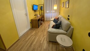 A hotel room with a wooden floor, a sofa, a small table, a TV, a desk, and a window with curtains. - NSM Hotel Cicolella (Foggia)