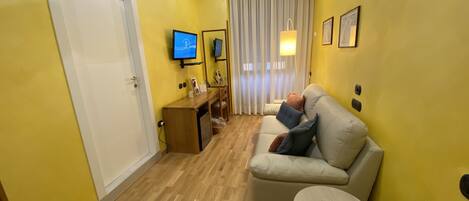 Habitación de hotel con suelo de madera, sofá, mesita, televisor, escritorio y ventana con cortinas.