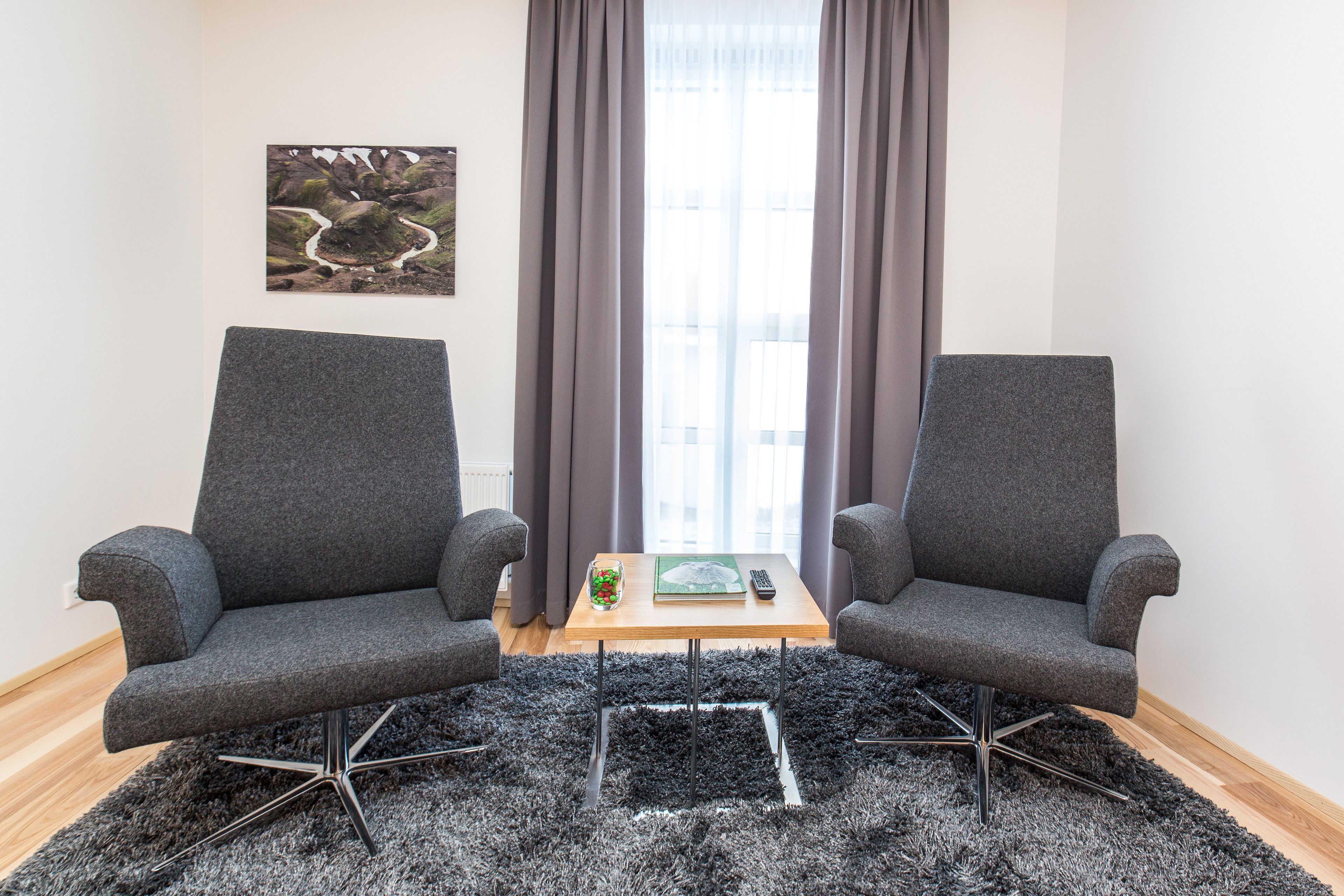 Una habitación con dos sillones grises enfrentados, una mesita de madera con un control remoto, un tarro de vidrio con aperitivos y un cuadro en la pared.