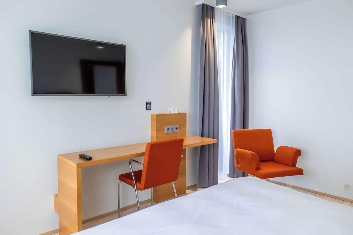 A hotel room with a bed, a desk with a chair, a TV mounted on the wall, and a window with curtains.