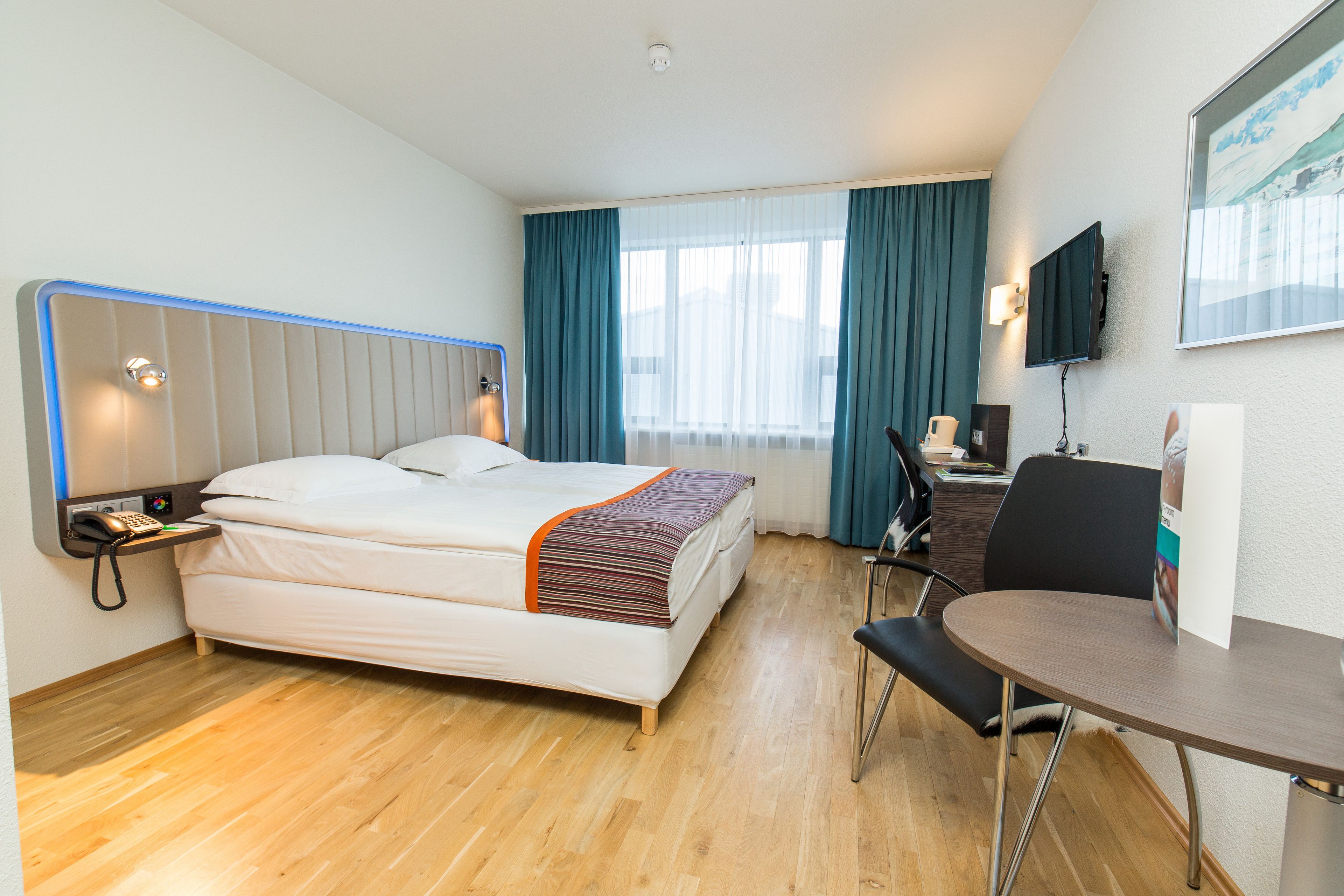 Et hotelværelse med seng, natbord, stol, skrivebord og fjernsyn.
