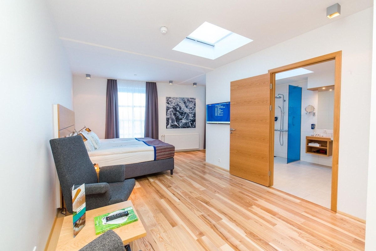 A modern hotel room with a bed, a desk, a chair, a TV, and a bathroom visible through an open door.