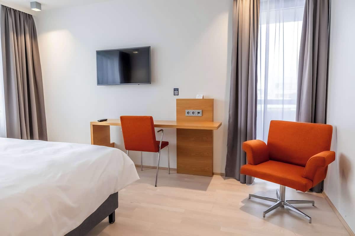 A hotel room with a bed, a desk with a chair, a TV mounted on the wall, and a window with curtains.