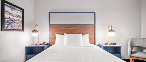 Habitación de hotel con una cama grande, dos mesitas de noche con lámparas, una silla y un cuadro en la pared.