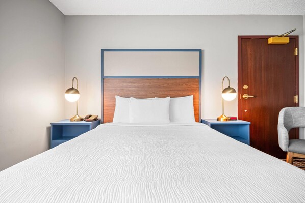 Una habitación de hotel con una cama grande, dos mesitas de noche con lámparas, un cabecero de madera, una puerta y una silla.